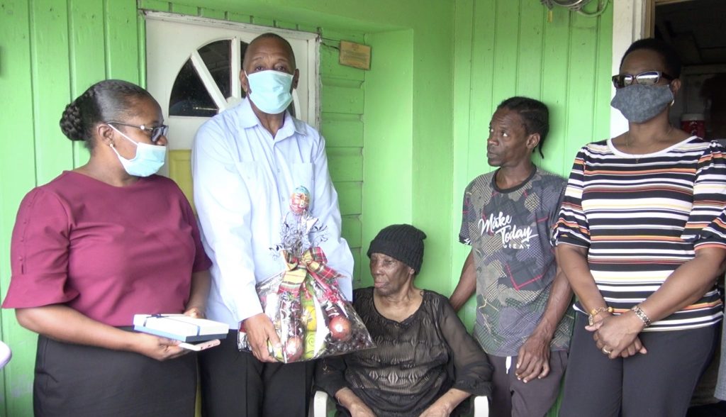 One hundred and one-year-old Eliza “Liza” Jeffers (sitting) with her nephew Randy at her side while Hon. Eric Evelyn, Minister responsible for seniors on Nevis, presents her with a fruit basket, a token of appreciation on behalf of the ministry and the Department of Social Services' Senior Citizens Division in celebration of National Centenarians Day on May 31, 2021
